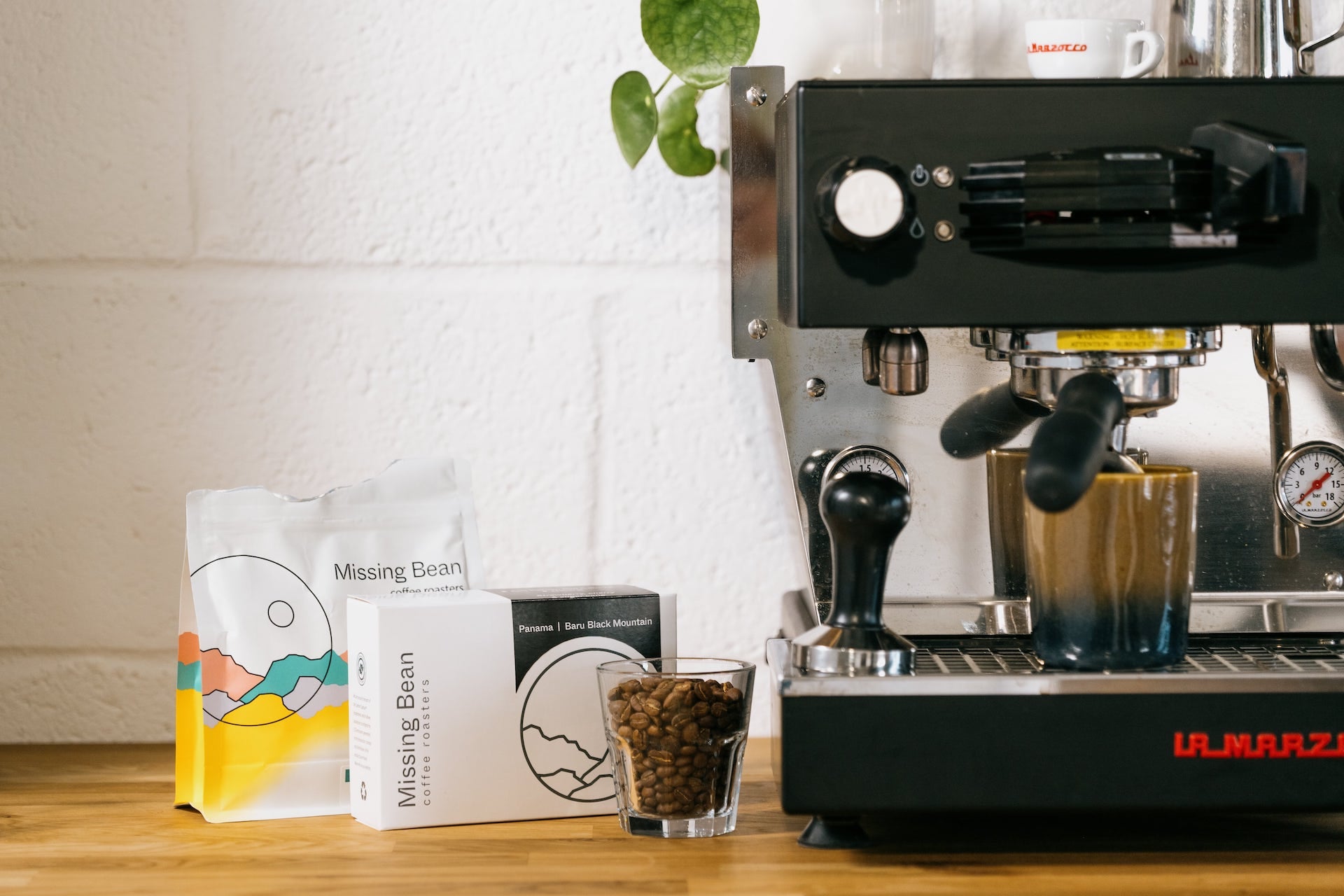 A coffee machine is placed on a wooden counter next to two Missing Bean coffee bean bags and a glass filled with coffee beans. A plant hangs on the wall in the background.