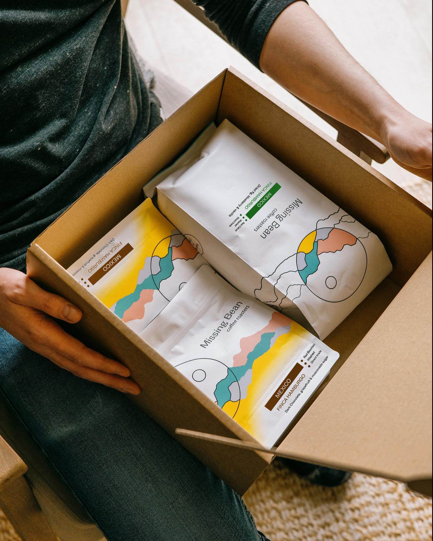 A man in blue jeans and a grey t-shirt opening a cardboard box filled with a variety of Missing Bean coffee beans