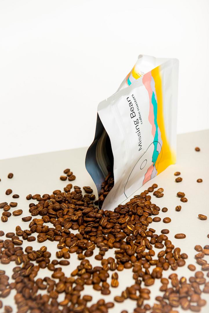 A 250g Missing Bean Coffee bag with freshly roasted coffee beans spilling on onto a white table.