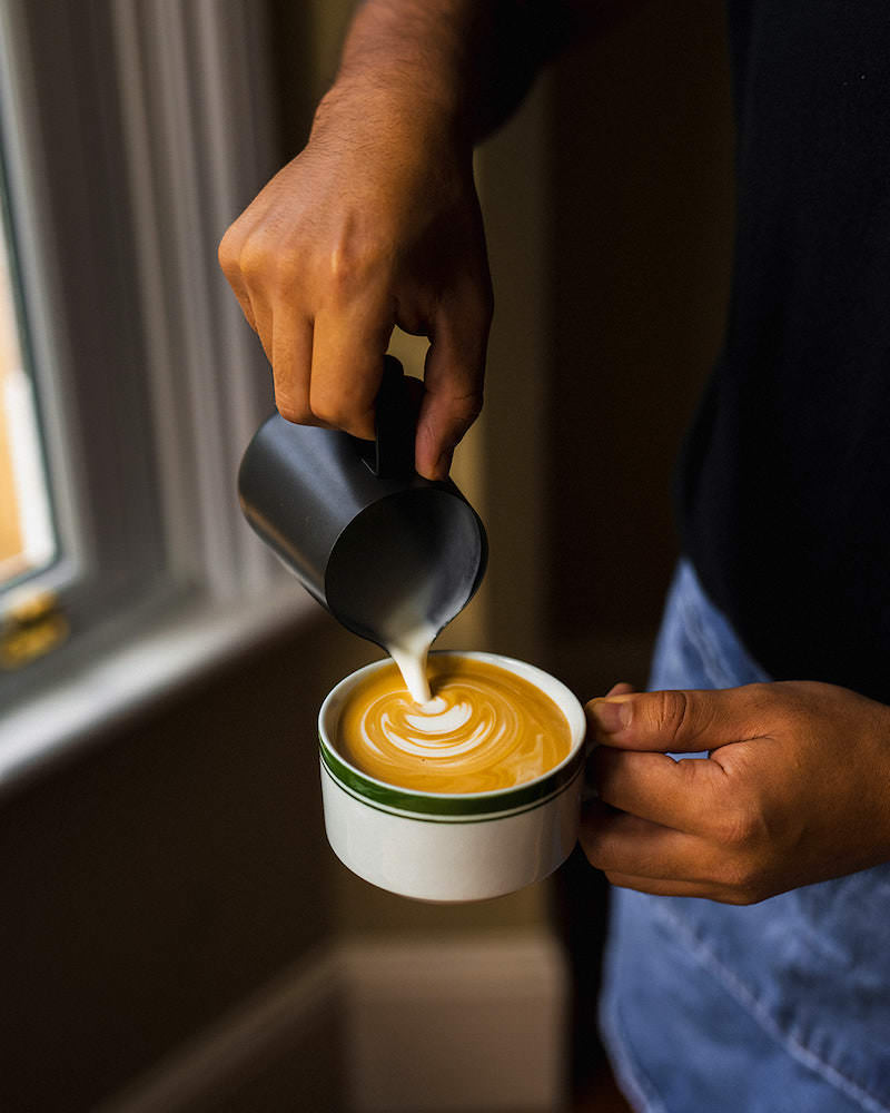 A person pouring milk into a coffee, made with Missing Bean coffee beans, against a window