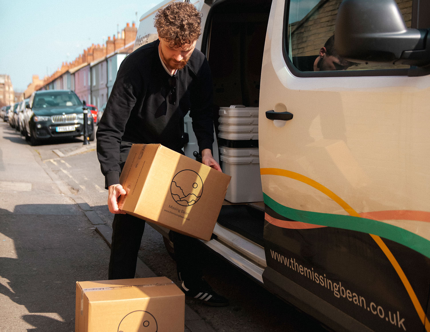 A male Missing Bean employee unloading cardboard boxes with Missing Bean logos out of the Missing Bean's branded elecrtic delivery band. The van is decorated with wavy stripes in Missing Bean colours, yellow pink and teal. In the van, you can see a stack of refillable coffee containers. The ban is parked on an Oxford street with colourful terraced houses in the distance. 