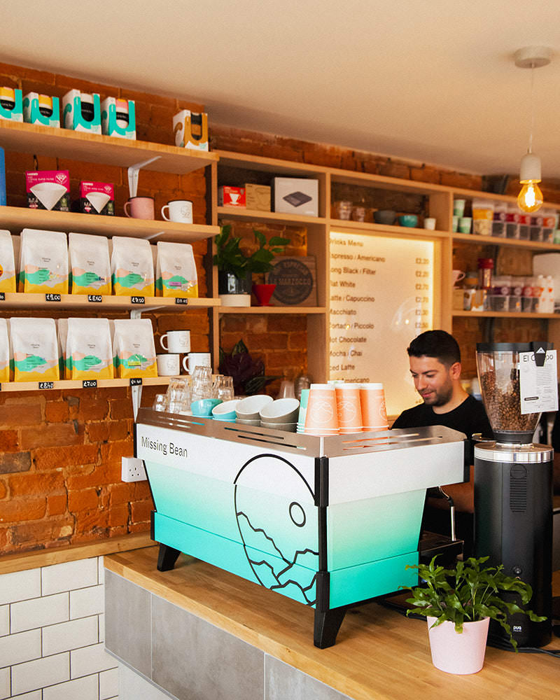 Barista behind counter of Missing Bean Banbury coffee shop, with custom blue gradient and logo La Marzocco coffee machine 