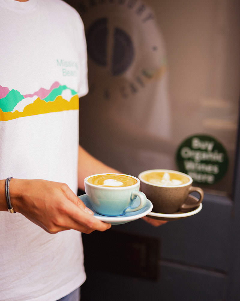 Charlbury Deli & Cafe barista wearing Missing Bean t-shirt delivering two lattes 