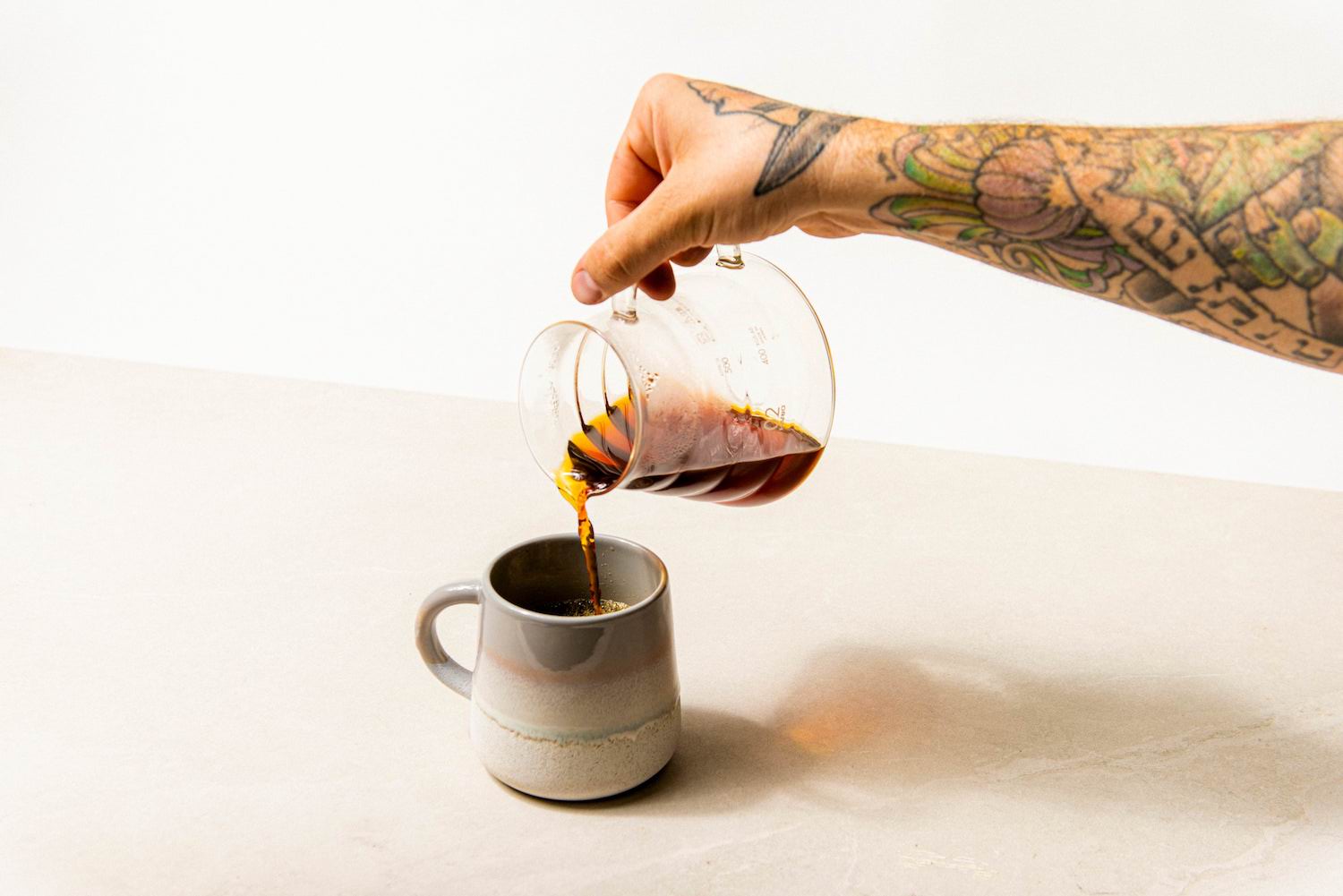  A man's tattooed arm pours black coffee from a glass Hario jar into a hand-thrown porcelain mug. The mug is predominantly grey with subtle pink and teal accents. The scene unfolds on a white marble table against a white wall.