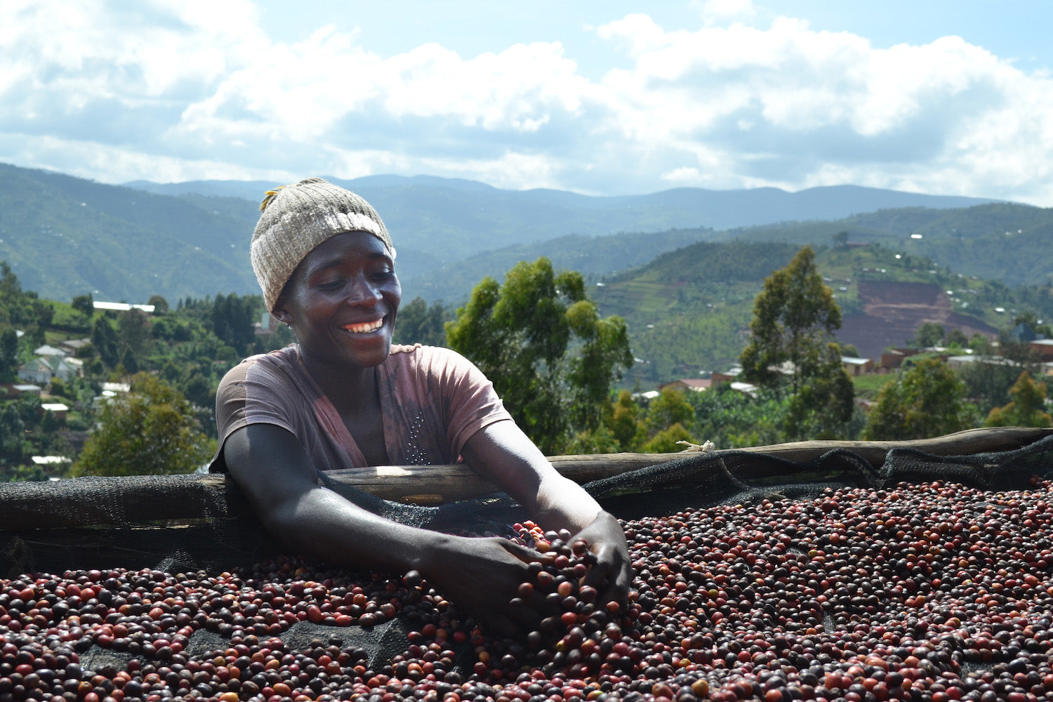 A female employee from the Migoti Coffee Company in Burundi, Africa. She is wearing a brown shirt and khaki beanie and smiling while washing coffee cherries at their washing station in the mountains. In the distance, you can see mountains and the town of Bujumbura. The sky is blue with scattered clouds. 