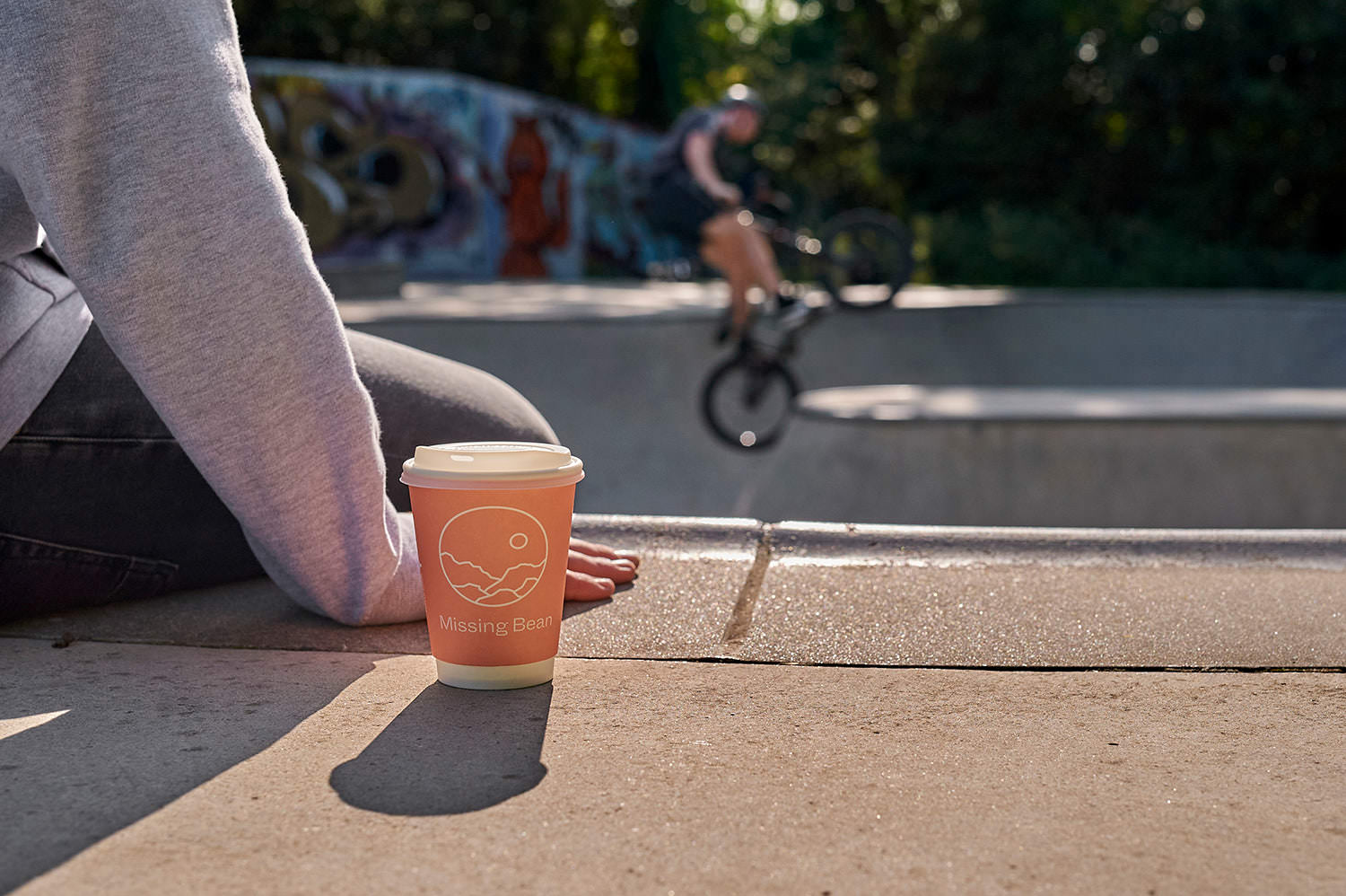 A person sitting at the edge of a skatepark bowl with a takeaway Missing Bean coffee watching someone doing a BMX trick in the distance