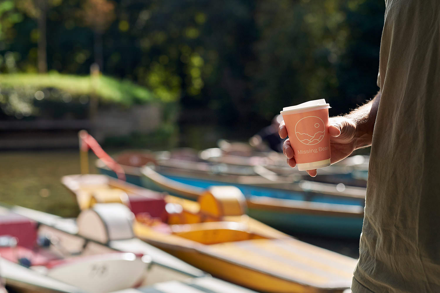 A man holding a pink Missing Bean takeaway coffee cup in front of rental boats on the Isis river in Oxford