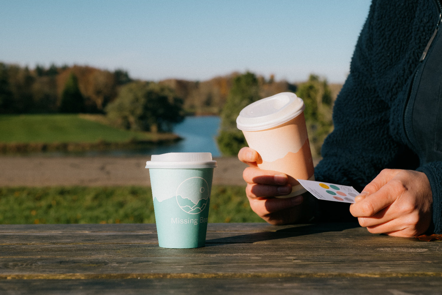 A person in a blue fleece, holding a Missing Bean loyalty card in one card, and a pink Missing Bean takeaway cup in the other. They are sat at a picnic bench with beautiful countryside scenes in the background. 