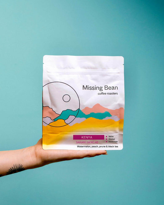 A 250g bag of Missing Bean Kenya Sakami Jane coffee beans, being held up in front of a blue background. 