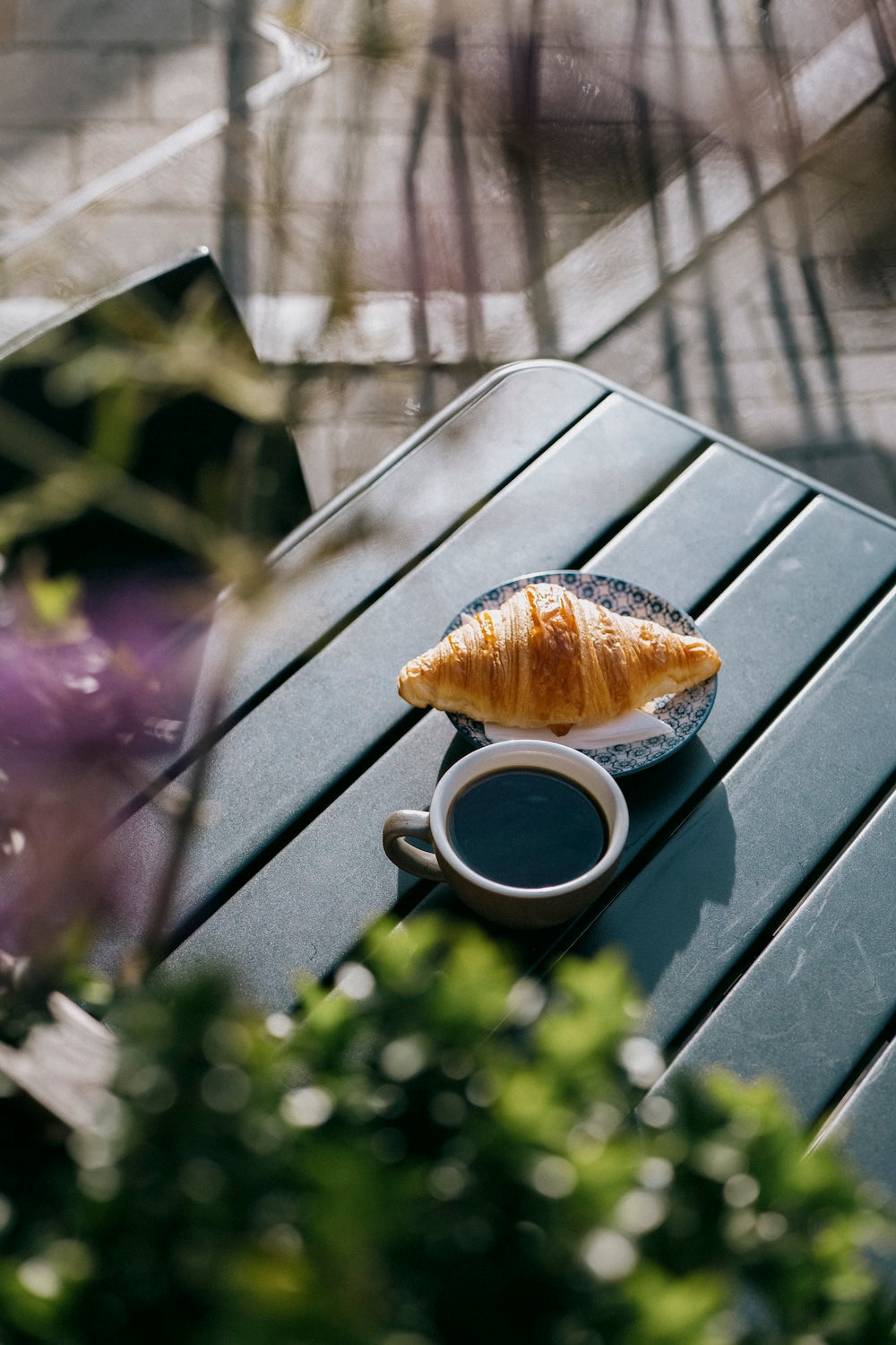 A croissant on a small plate and a cup of black coffee on a dark gray slatted table, photographed from above with soft-focus greenery in the foreground at Missing Bean's Botley cafe.