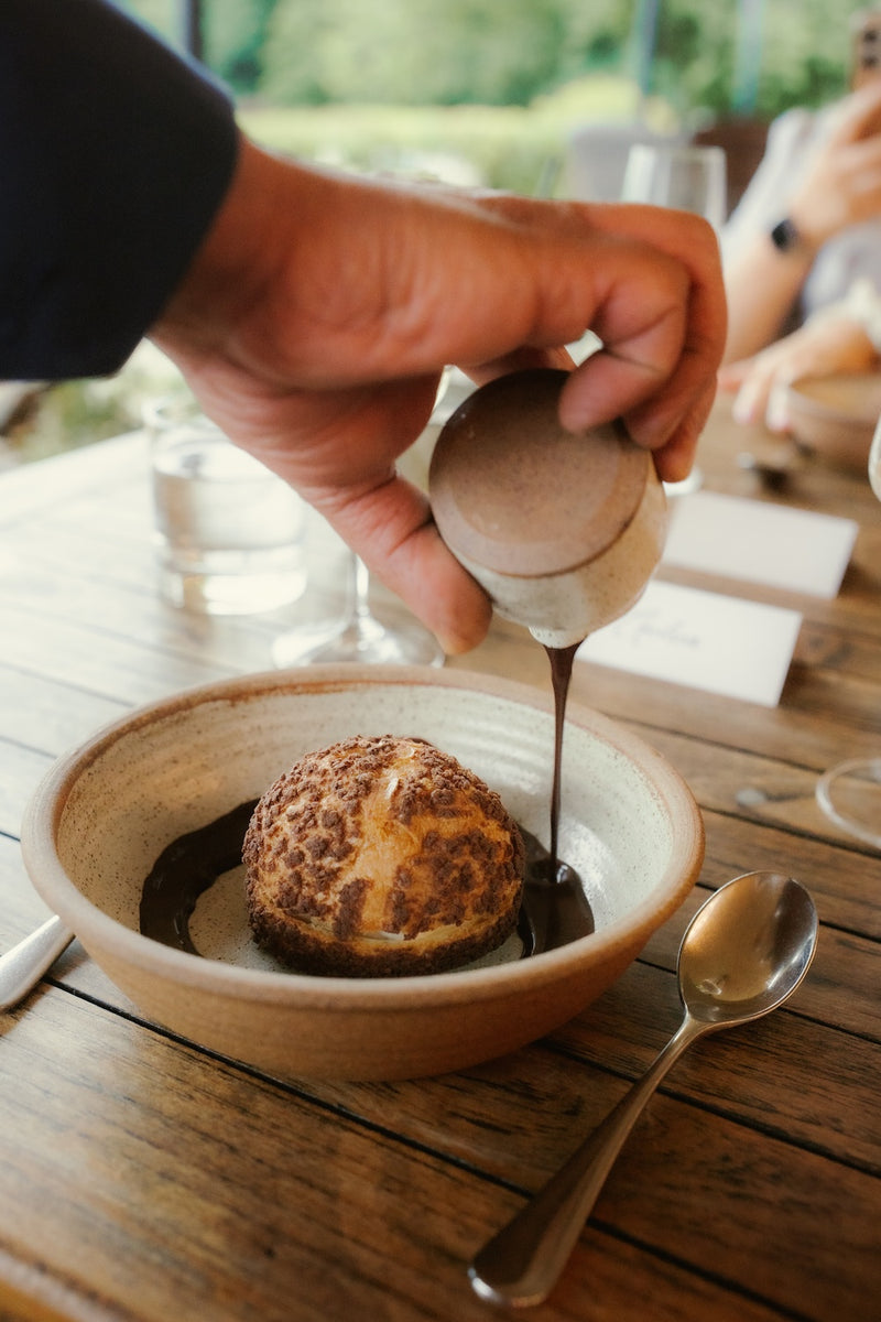 A close-up of a hand pouring dark chocolate sauce from a small ceramic pitcher into a bowl containing a tirami-choux crafted with Missing Bean coffee creme patissiere on a rustic wooden table at The Feathered Nest Inn in the Cotswolds.