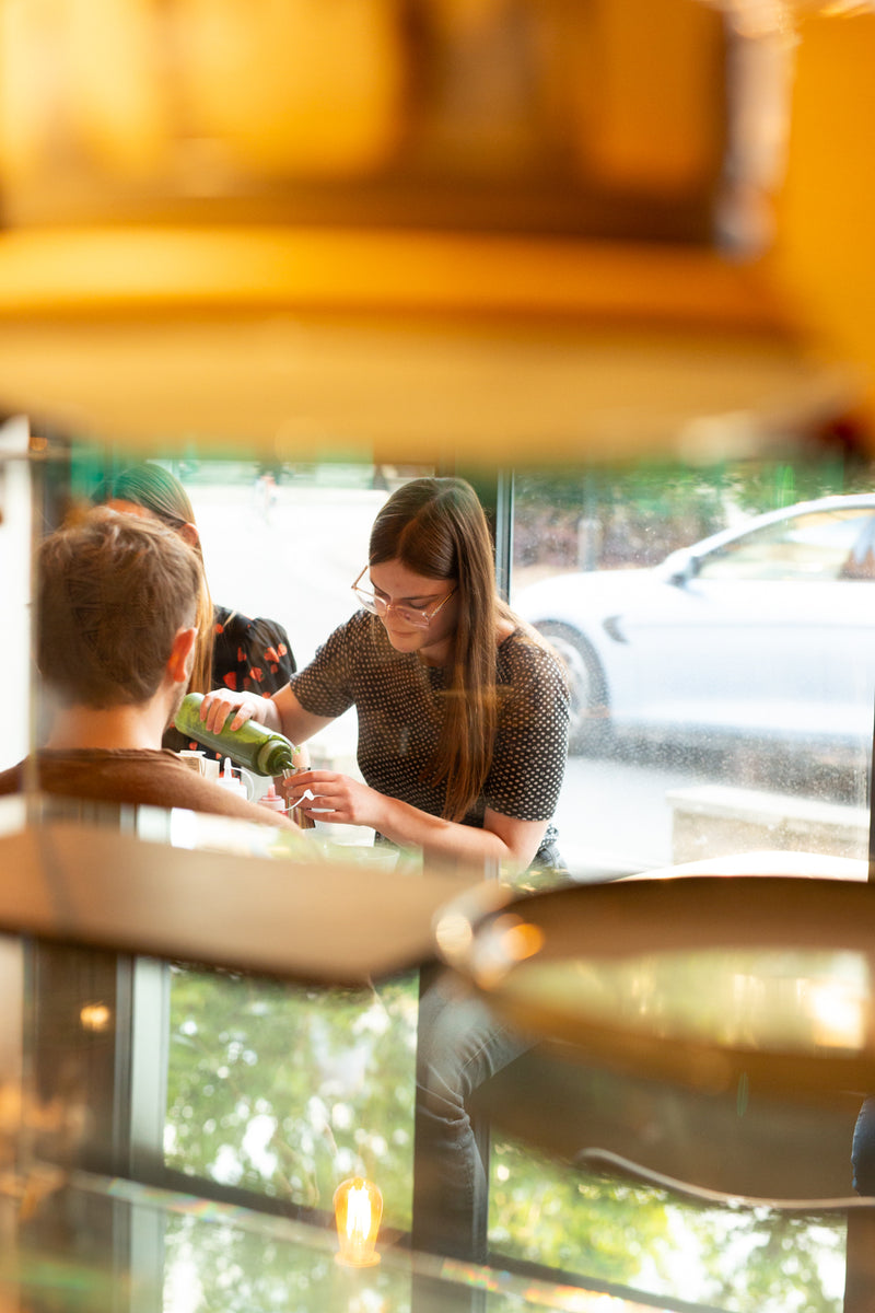 A woman in Missing Bean's Abingdon Cafe pours matcha into a glass during Missing Bean's Matcha Masterclass, viewed through the counter with reflections and blurred foreground elements.