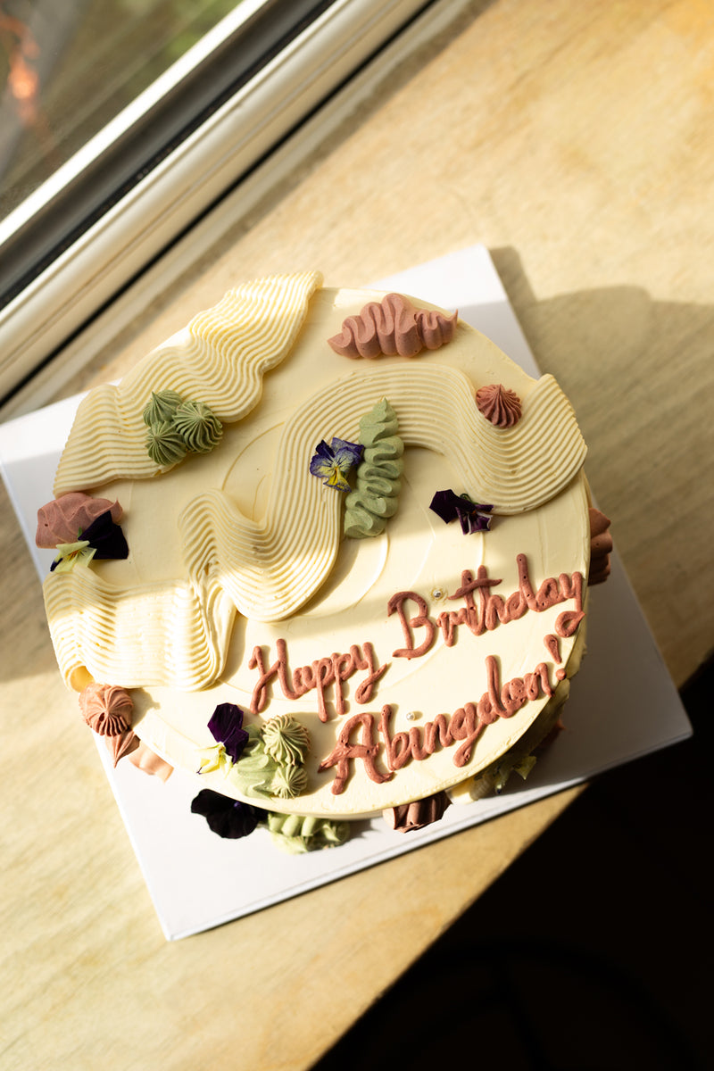A round birthday cake, for Missing Bean Abingdon's 5th birthday celebration, with cream-coloured frosting and piped decorative swirls, placed on a white square board on a wooden table near a window. The cake is decorated with small edible flowers and has 'Happy Birthday Abingdon!' written in pink icing.