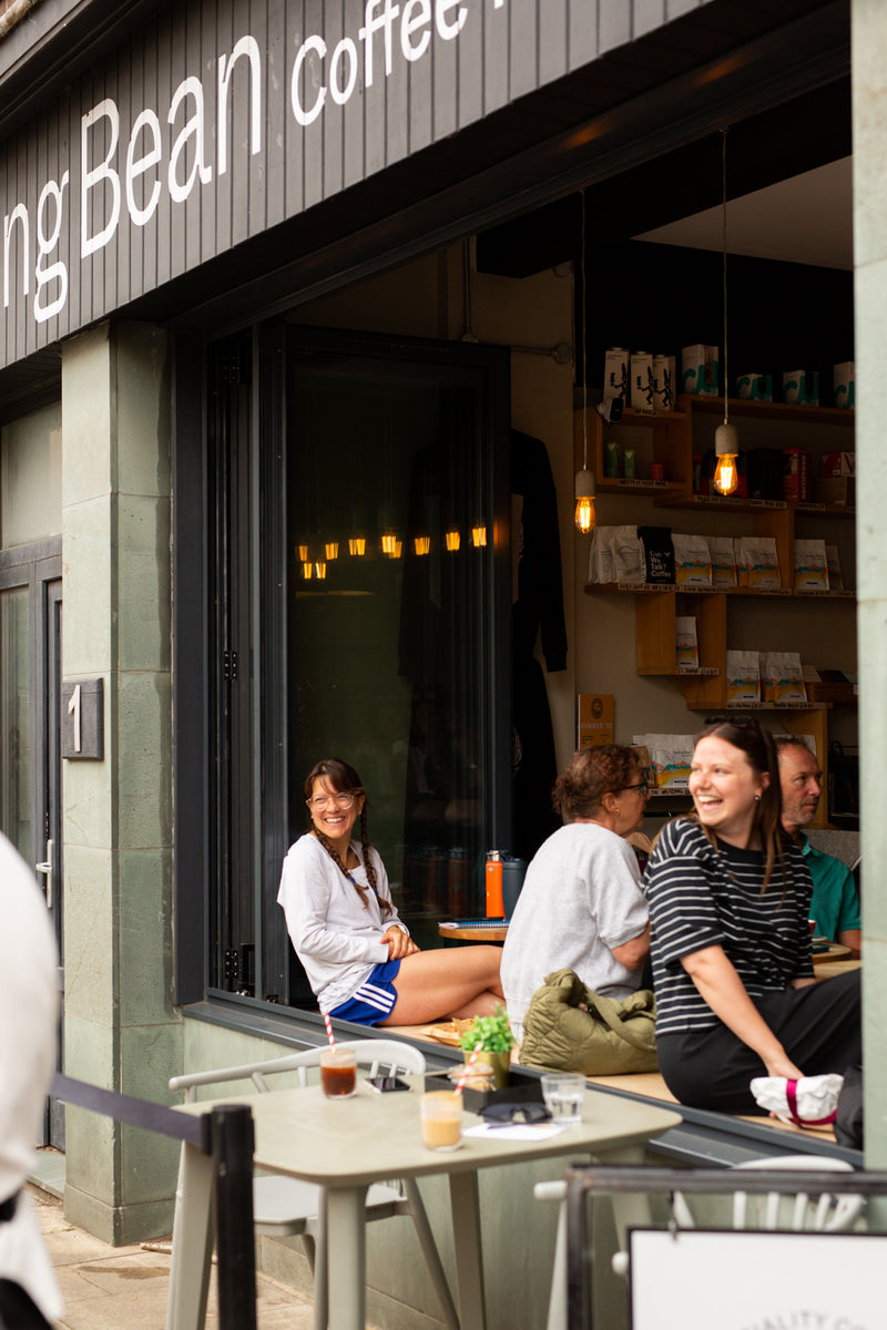 Several people sit and relax outside Missing bean's Abingdon coffee shop with a large window and outdoor seating. The shop sign reads 'Missing Bean Coffee Roasters', and shelves with Missing Bean coffee are visible inside.
