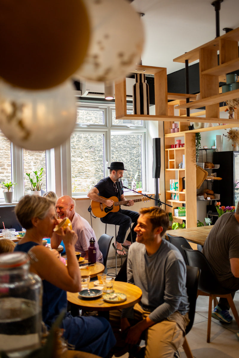 Missing Bean's Abingdon cafe during their 5th Birthday celebrations with several people seated at small round tables, while musician Ear Candy Oxford in the background plays an acoustic guitar. Large decorative balloons are partially visible in the foreground, and wooden shelving and windows frame the scene.