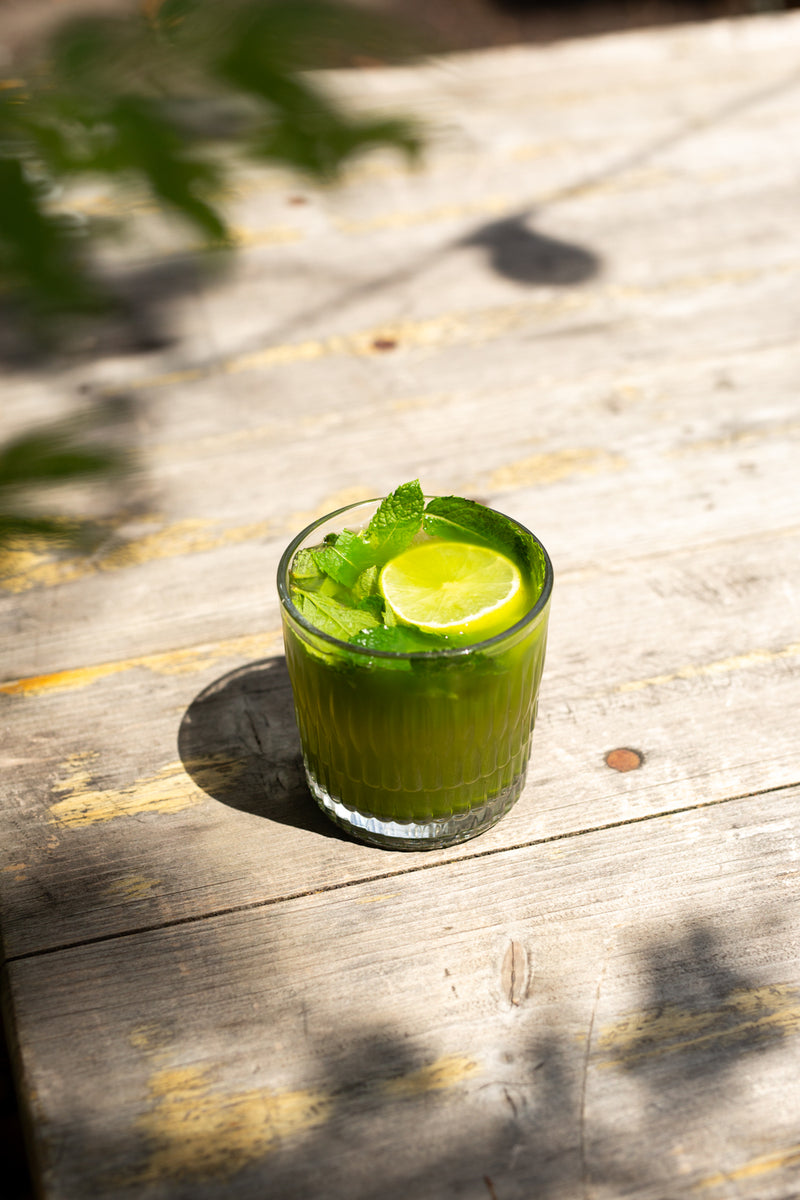A clear glass tumbler filled with Missing Bean's Summer Special, the Matcha Mojito, garnished with a lime slice and mint leaves, set on a weathered wooden surface with dappled sunlight and shadows.