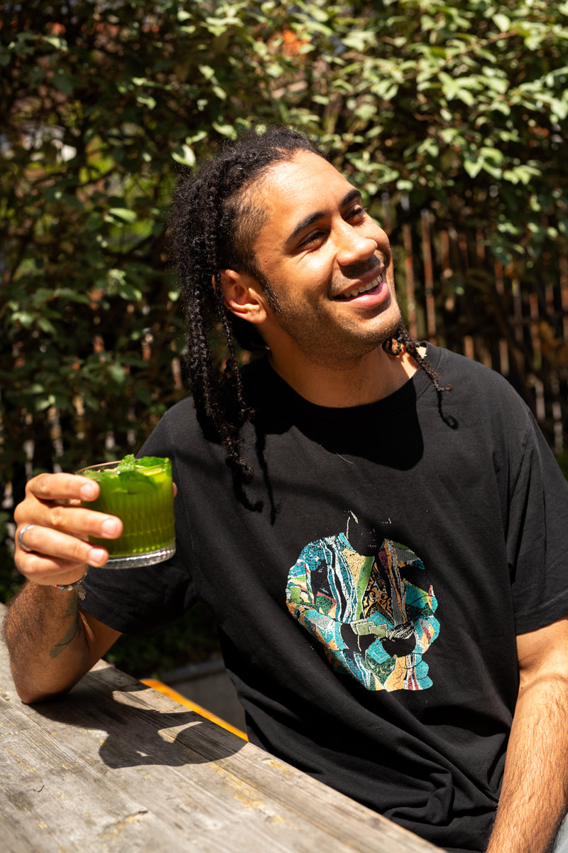 Missing Bean's Head Roaster, Brian, sits outdoors on a wooden table, smiling and holding a Missing Bean's Matcha Mojito, and green drink with mint leaves and limes visible. He is wearing a black t-shirt with a colourful graphic print on the front.