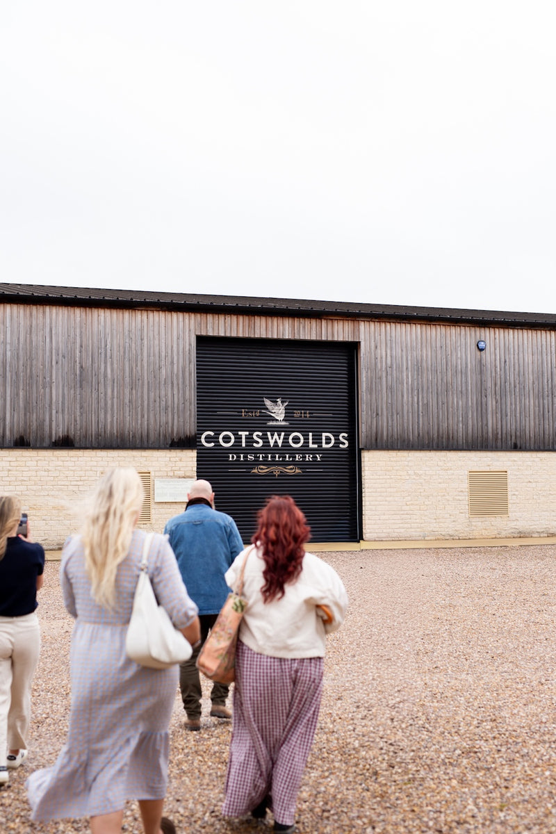 Several people walk toward a large Cotswolds Distillery building with a dark garage-style door and wood siding under an overcast sky.
