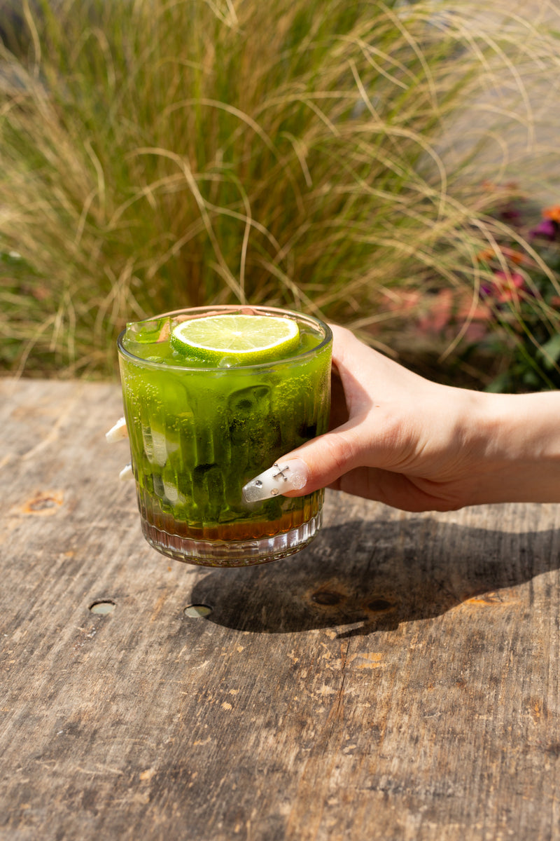 A hand with long white manicured nails holds a clear glass tumbler filled with Missing Bean's Summer Special, the Matcha Mojito, a green drink with lots of ice and a lime wheel, set on a rustic wooden table at Missing Bean's Roastery Cafe with long grass and flowers in the background.