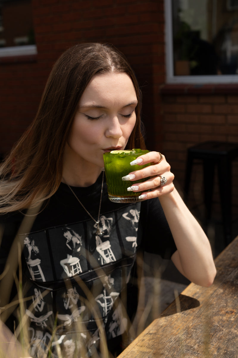 A woman with long brown hair is outside Missing Bean's Roastery Cafe, drinking from a glass tumbler, full of Missing Bean's Summer Special, the Matcha Mojito. A green drink with a lime wheel, ice and mint leaves. She is wearing a black graphic t-shirt with white illustrations.