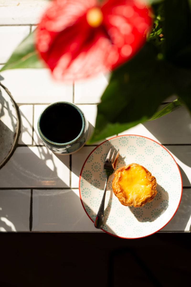 A top-down view of a breakfast table scene with a Portuguese tart on a patterned plate, a blue mug of batch brew, and a blurred red flower in the foreground on a white tiled surface.