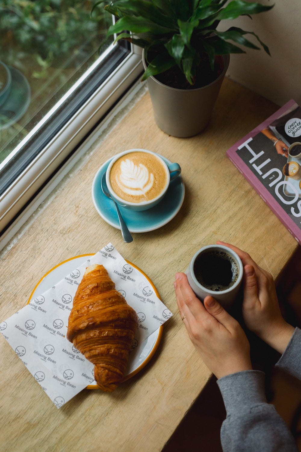 A cozy cafe table scene at Missing Bean Abingdon with a croissant on Missing Bean branded paper, a latte with latte art, another cup of black coffee held by hands, and a potted plant near a window.