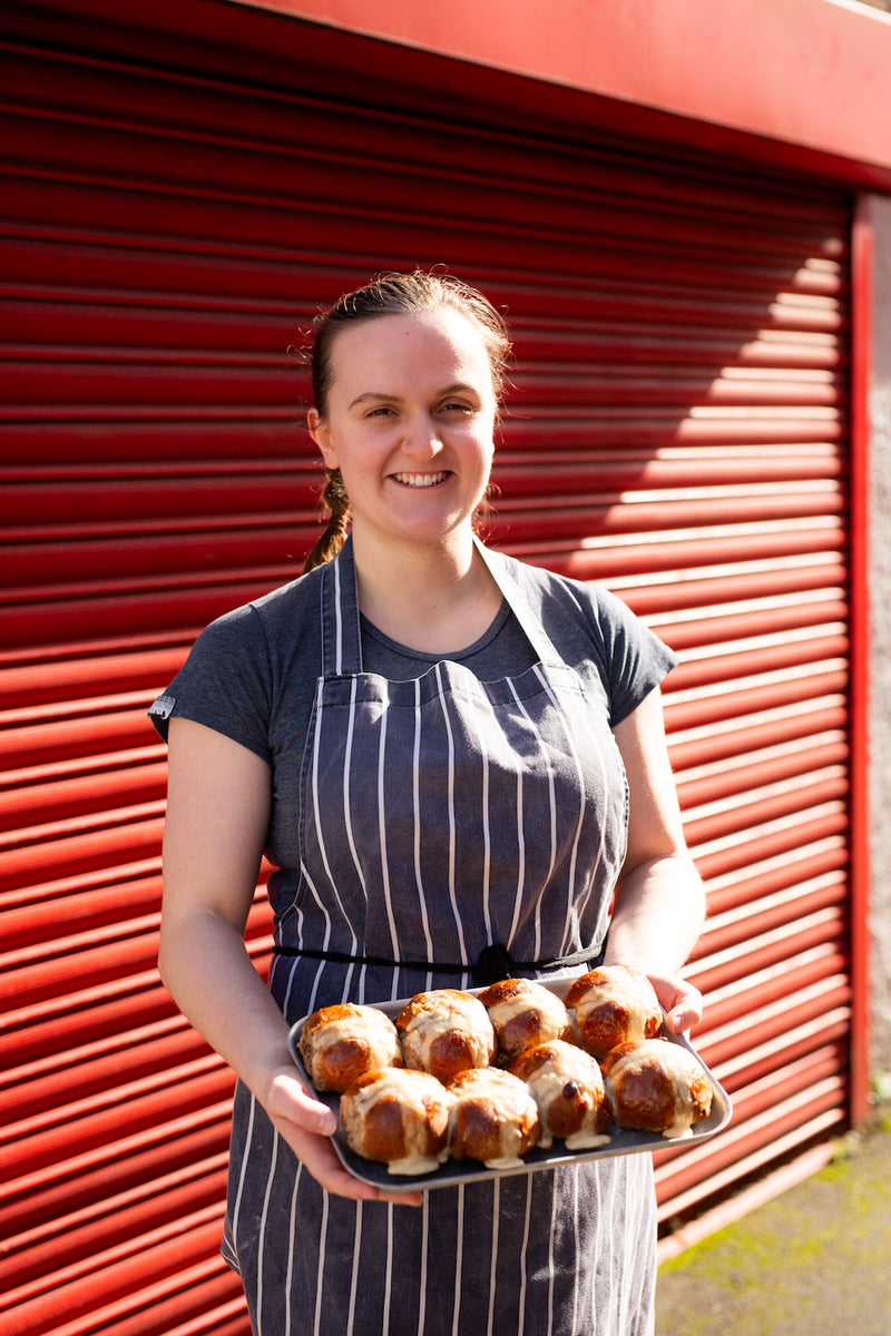 Missing Bean baker, Laura, holding a tray of hot cross buns in front of a red shutter