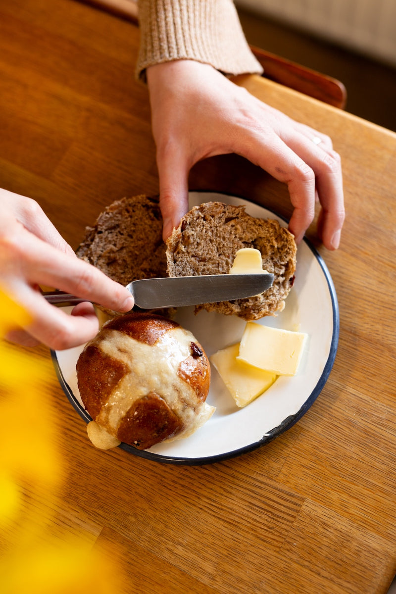 Person spreading butter on a hot cross bun with a knife on a plate, on a wooden table.