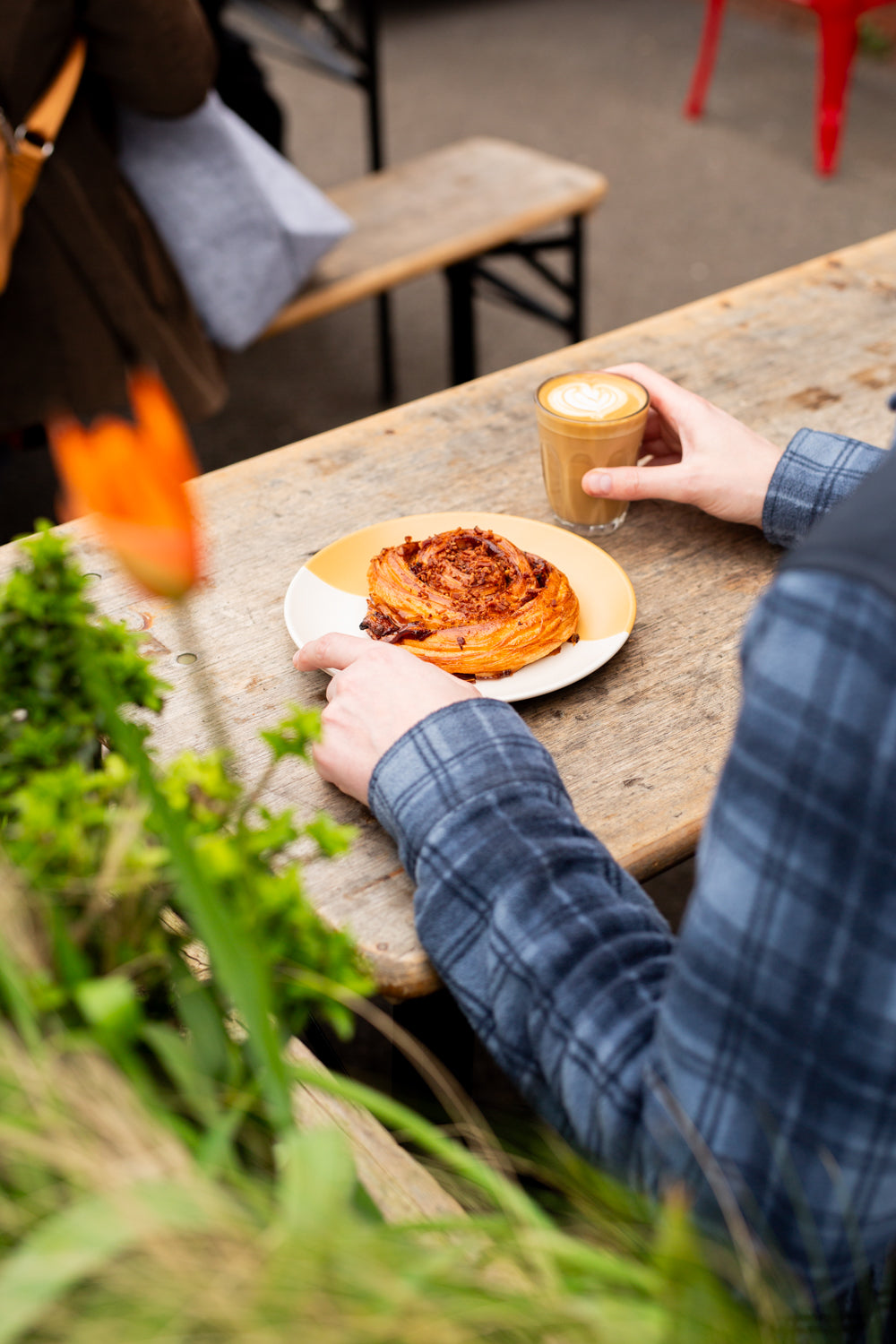 A person sitting at a rustic wooden cafe table outside Missing Bean's East Oxford cafe, holding a plate with a miso maple bacon swirl pastry and a latte in a glass. The scene is framed by green plants in the foreground and a wooden bench in the background.