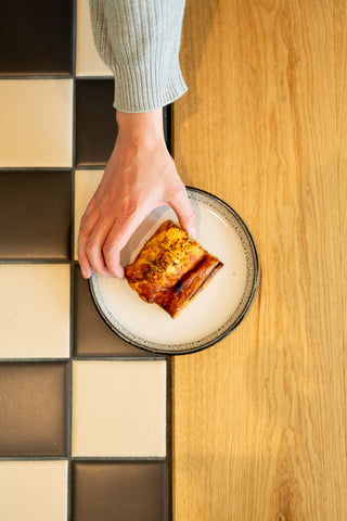 Person holding a sausage roll on a half wooden half black and white tiled surface.