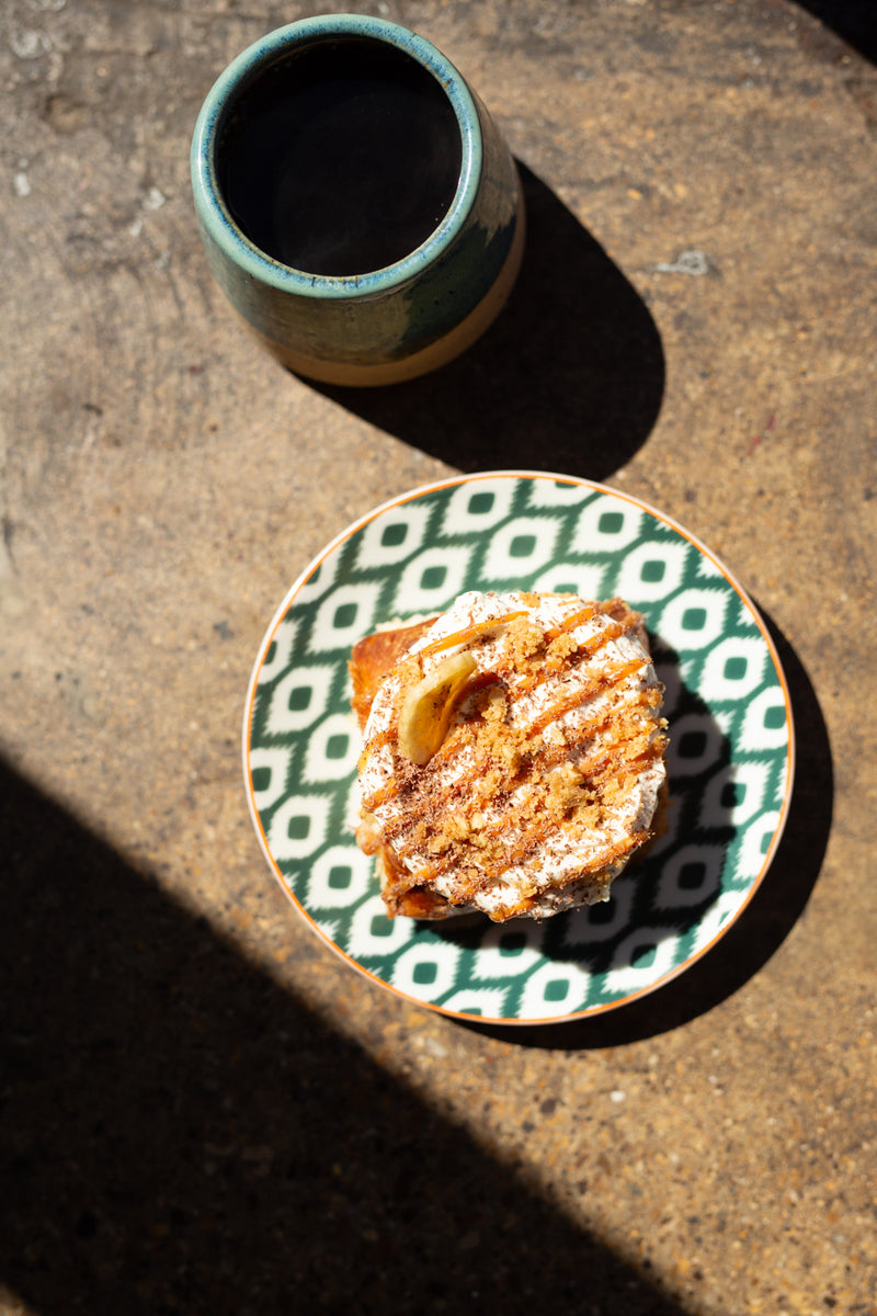 A top-down view of a banoffee batch bun on a green and white patterned ceramic plate, next to a matching ceramic mug filled with batch brew on a textured stone surface with strong sunlight and shadows.