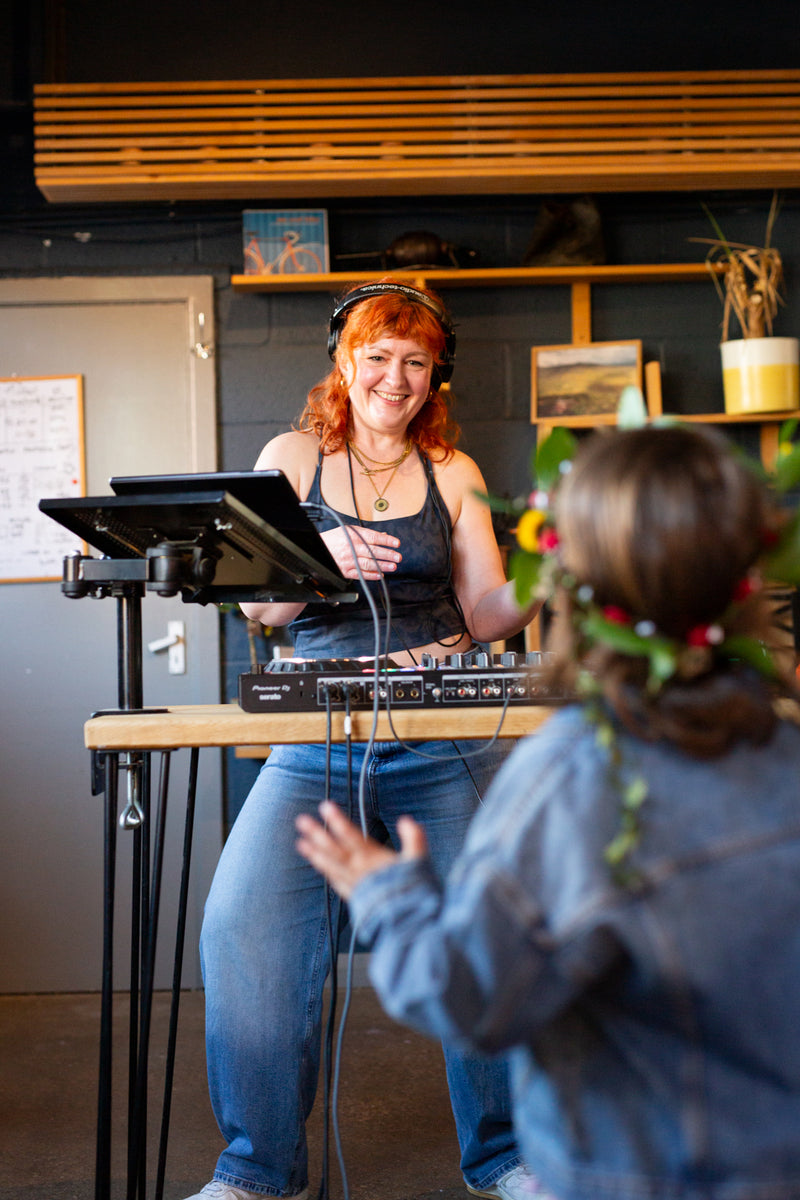 The DJ Rosie Raves, with red hair wearing headphones and a tank top stands behind a DJ booth mixer, smiling while a child in the foreground dances. The setting is Missing Bean's Roastery cafe with wooden shelves and plants.