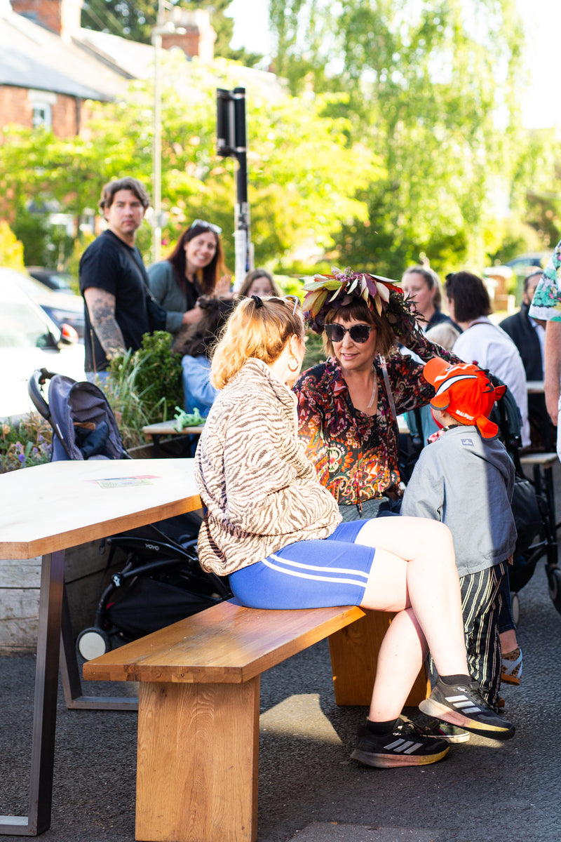 A woman in a tiger patterned top and blue shorts sits on a wooden outdoor bench at Missing Bean's Roastery Cafe, surrounded by other people, tables, and greenery in the background for Missing Bean's May Morning Rave.