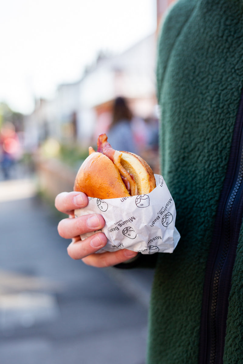 A person in a dark green fleece jacket holds a bacon roll in Missing Bean branded paper on a blurred street background.