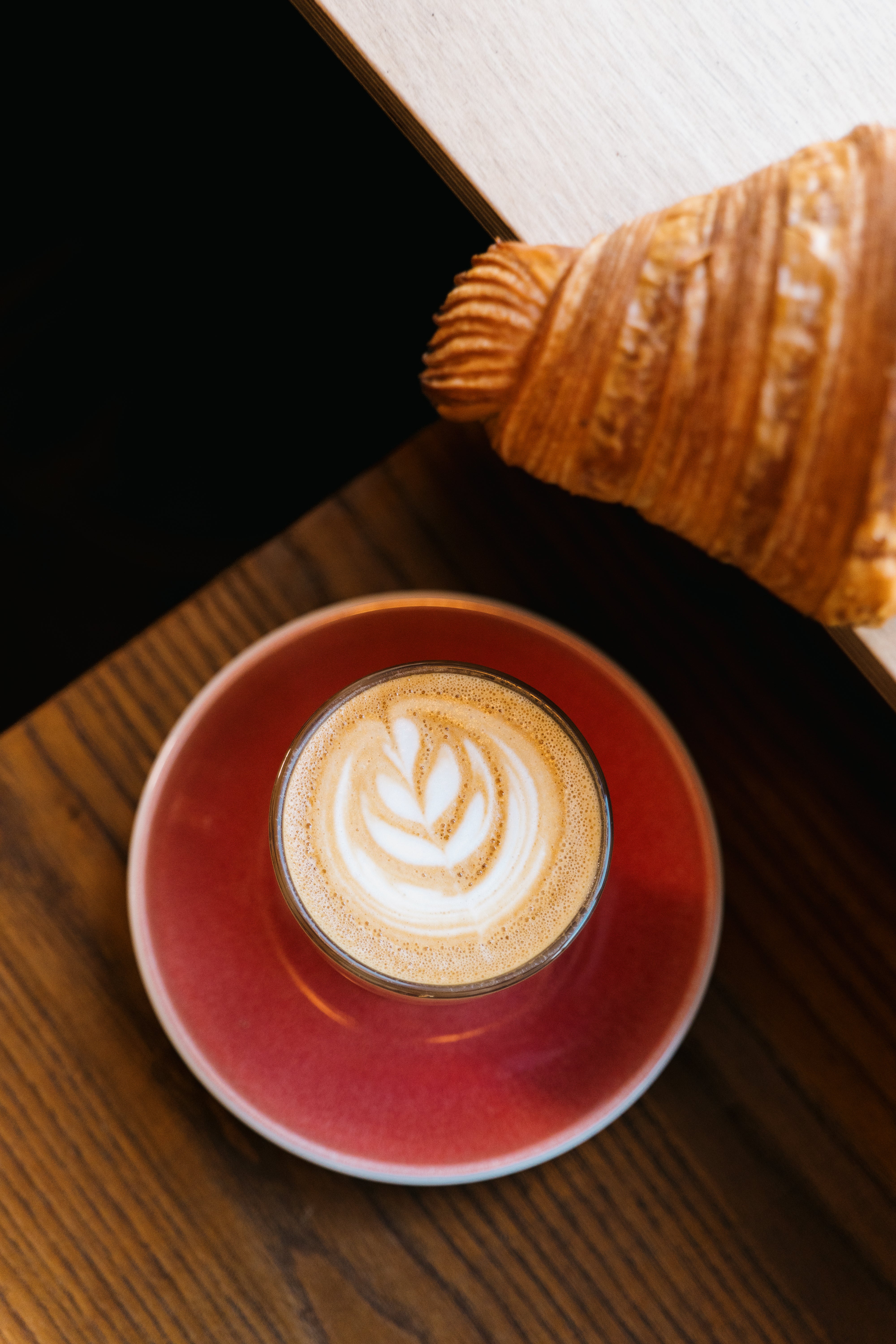 Cup of coffee with latte art on a red saucer next to a croissant on a wooden surface.