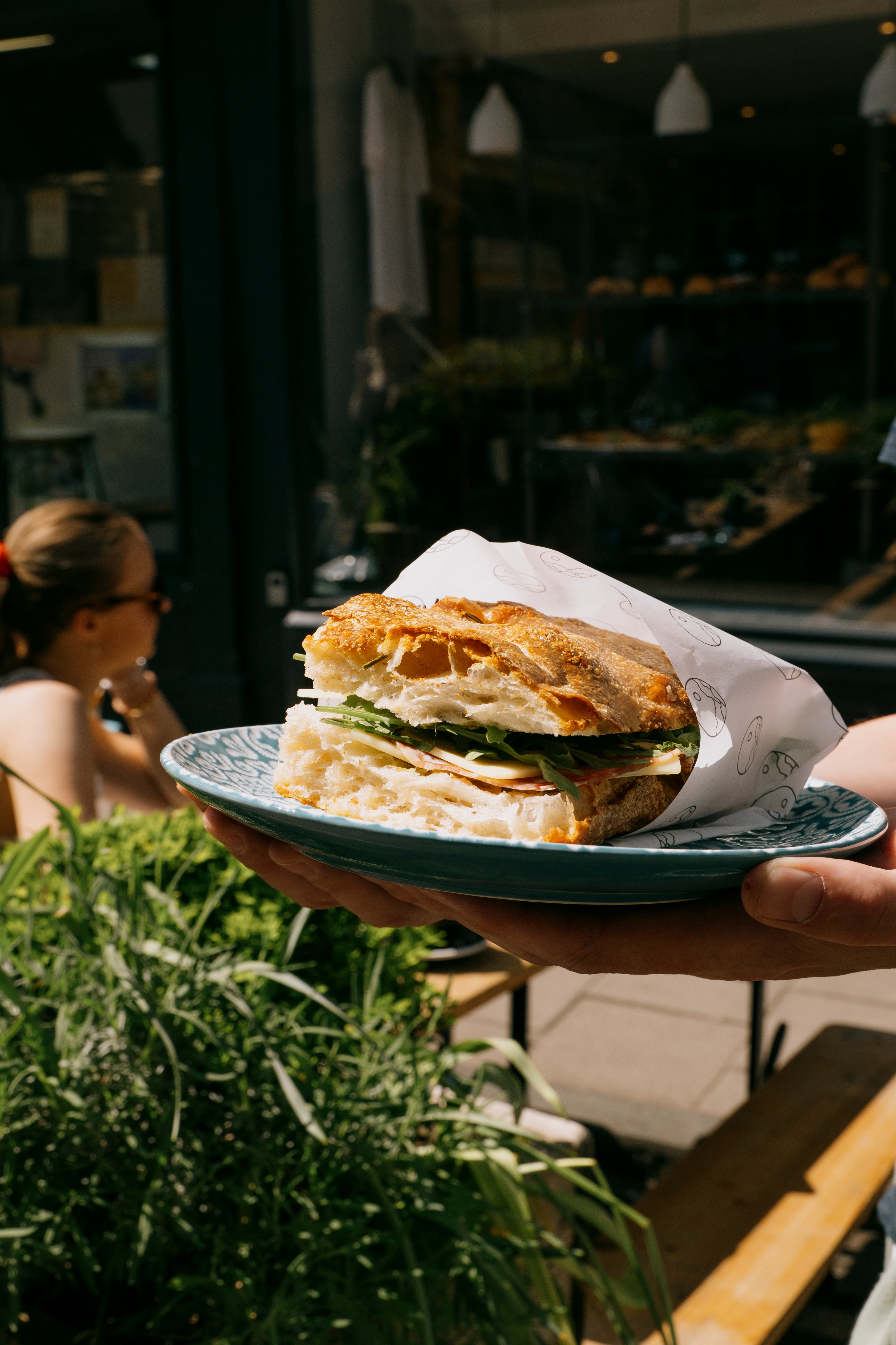 Person holding a plate with a sandwich outside a Missing Bean coffee shop