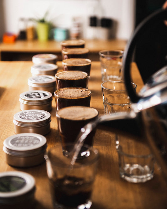 Coffee cupping process with coffee grounds, tins, and a glass on a wooden surface.