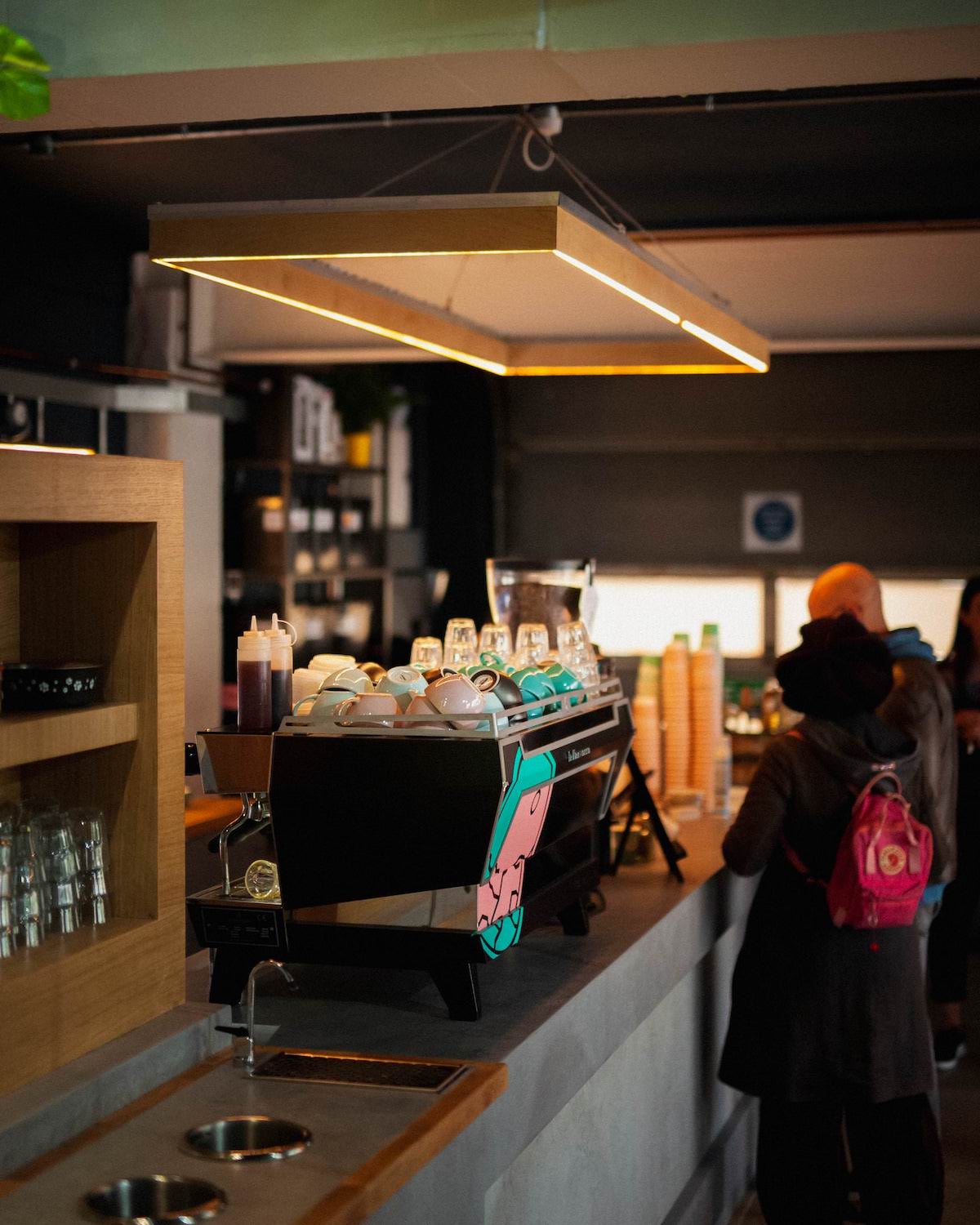 Customers at Missing Bean East Oxford Roastery, in front of custom La Marzocco coffee machine with missing bean logo in teal and pink