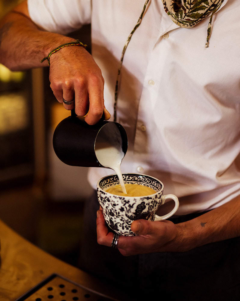 Thyme barista, wearing a white shirt and scarf, pouring a coffee made using Missing Bean coffee into a black and white china teacup