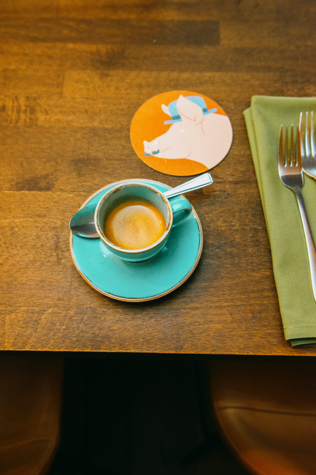 Teal mug and saucer, filled with Missing Bean wholesale coffee, next to a Three Little Pigs Wallingford beermat on a wooden table with two forks on a green napkin