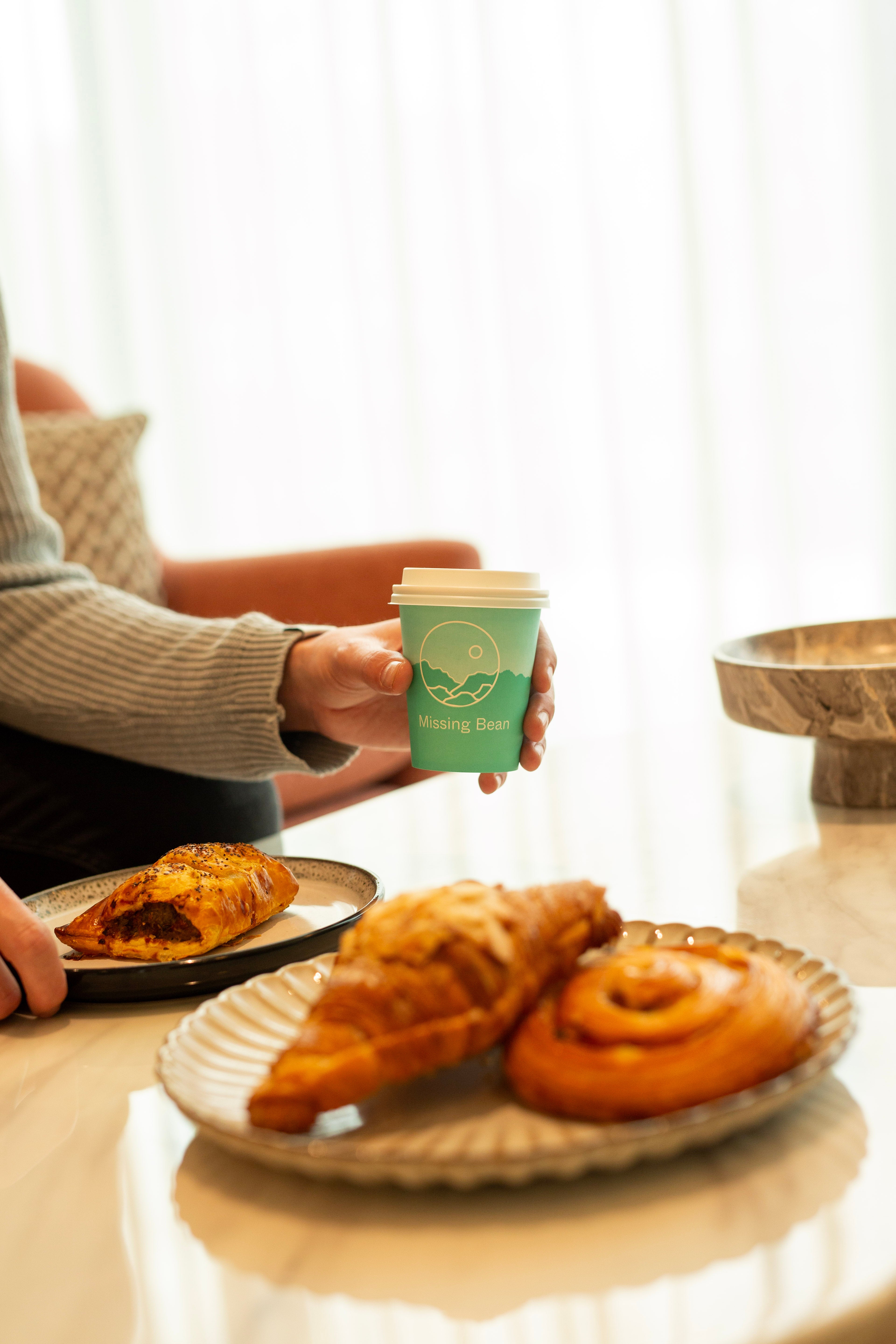 Person holding a Missing Bean takeaway coffee cup with a sausage roll, almond croissant and pain au raisin on a marble table.