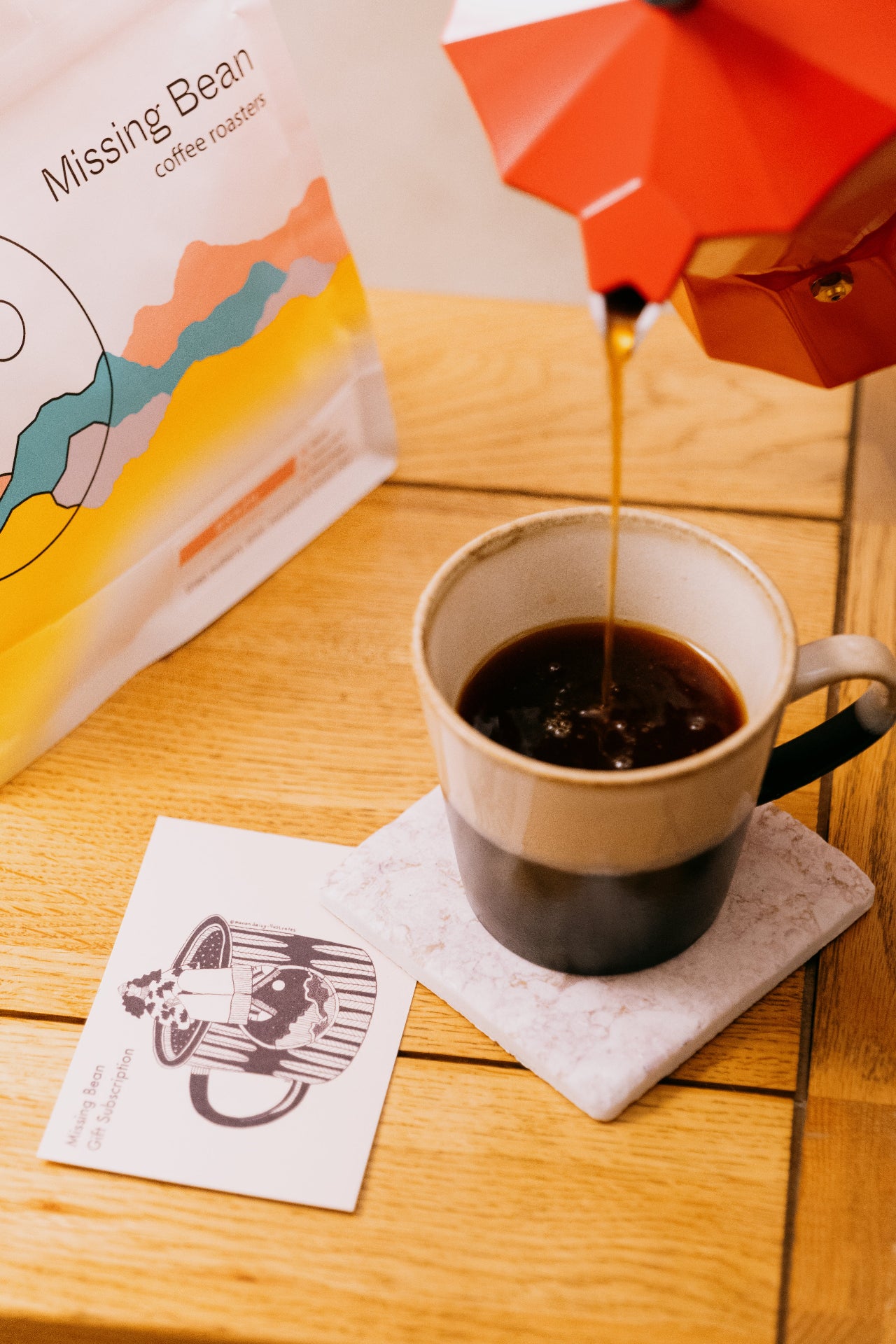 A red cafetiere pours Missing Bean's coffee into a white and grey mug. A Missing Bean gift subscription card can be seen on the table. 
