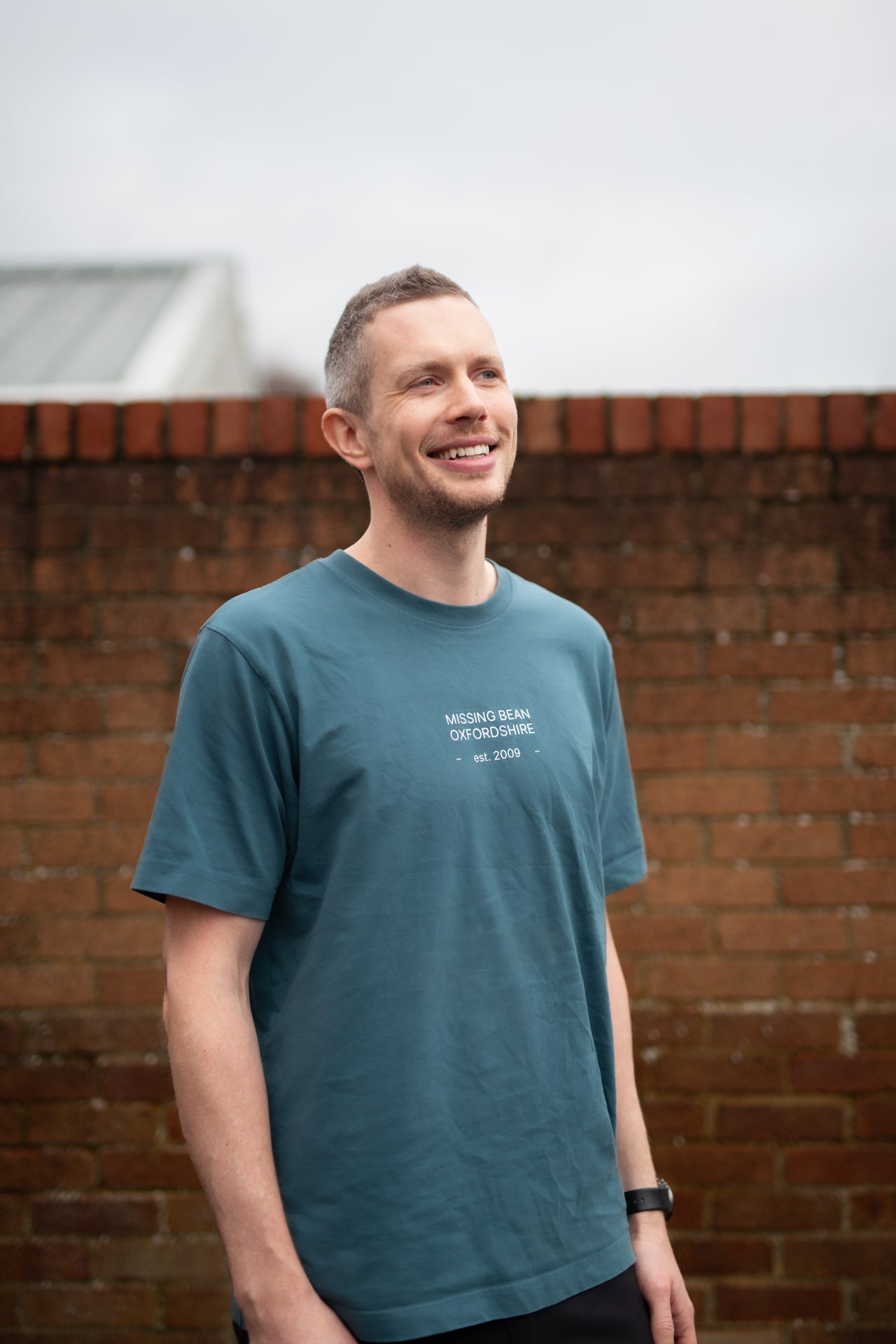 Coffee Support & Roastery Assistant, Nic Kerrigan, smiling  wearing a blue Missing Bean t-shirt in front of a brick wall. 