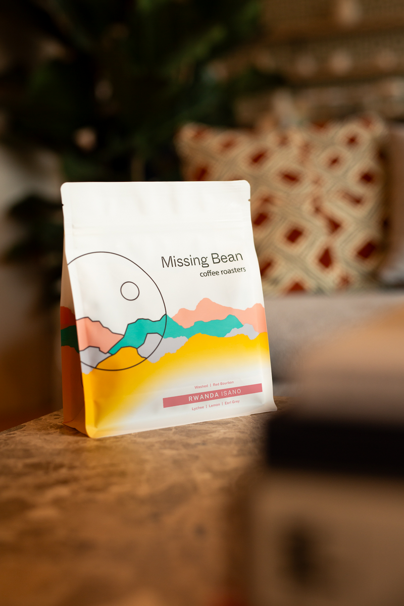 A bag of Missing Bean's Rwanda Isano coffee beans, standing on a wooden surface with a blurred indoor background.