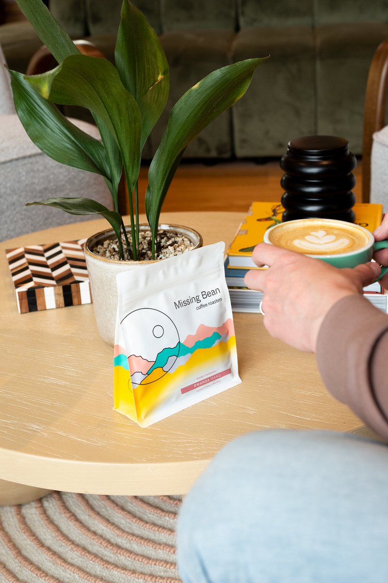 A person holding a cup of latte art coffee sits at a light wood table next to a potted plant and a bag of Missing Bean's Rwanda Isano coffee.