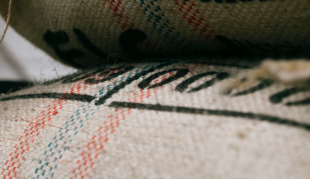  A close-up of a hessian sack, housing the globally-sourced coffee beans by Missing Bean. The text on the sack, rendered in stenciled black font, is too close to discern clearly but seems to consist of numbers. Additionally, the sack features embroidered stripes—a red one, followed by a blue one, and another red stripe.