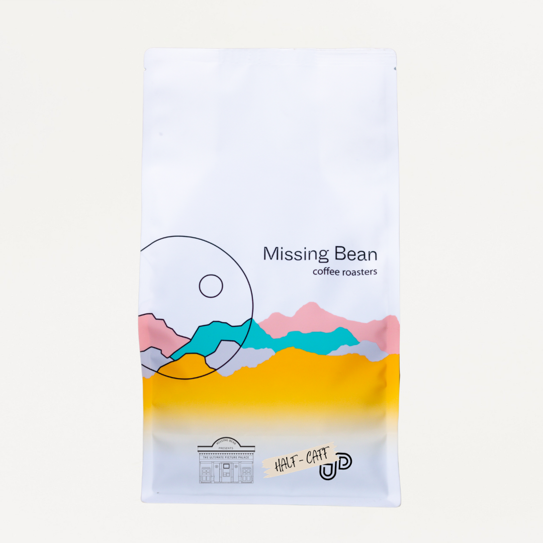 A white bag of Missing Bean's Ultimate Picture Palace coffee, featuring a colourful graphic of stylised mountains in pink, teal, yellow, and gray, with 'HALF-CFF UPP' written at the bottom.