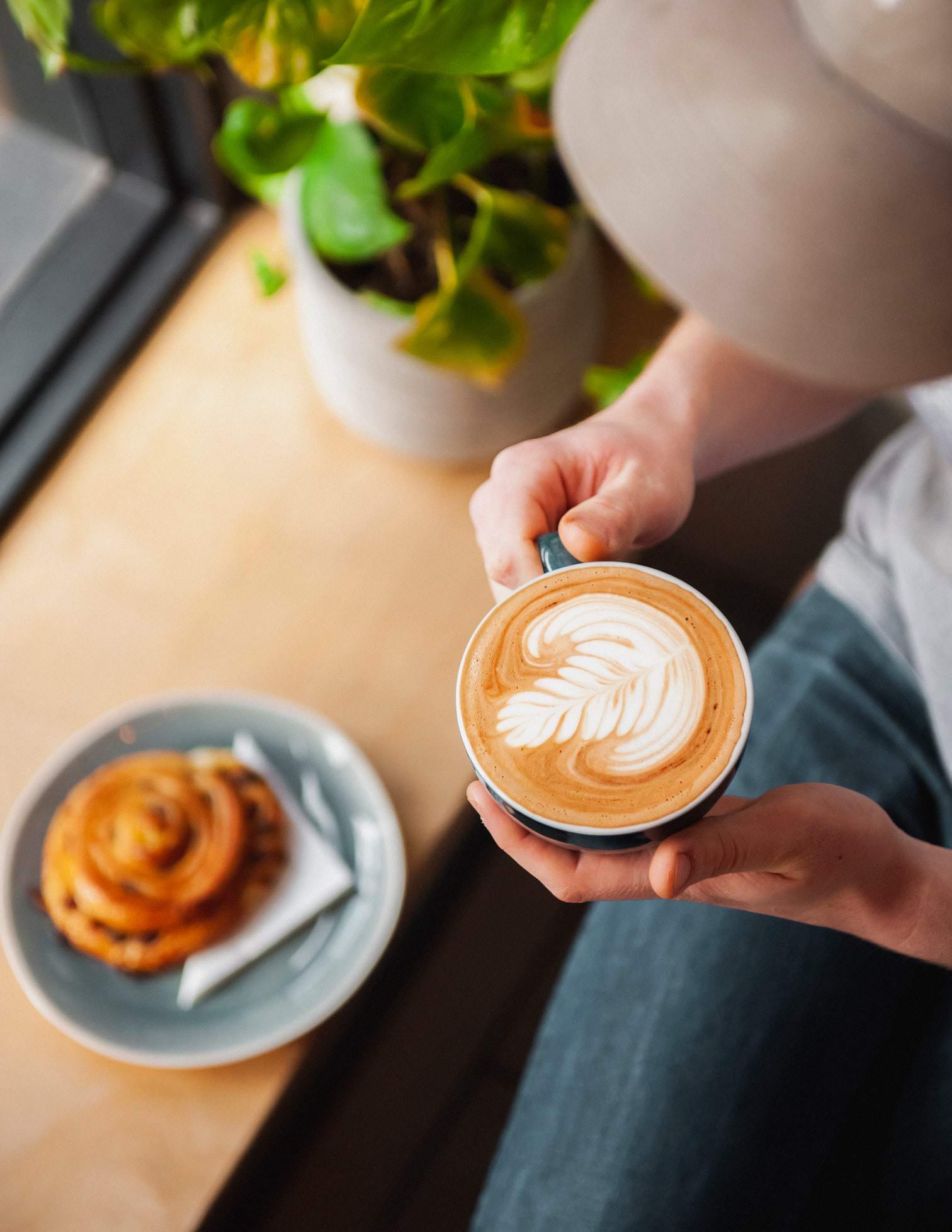 A person holding a cappuccino at a table with a pastry and a plant in the window of the Missing Bean coffee shop in Abingdon