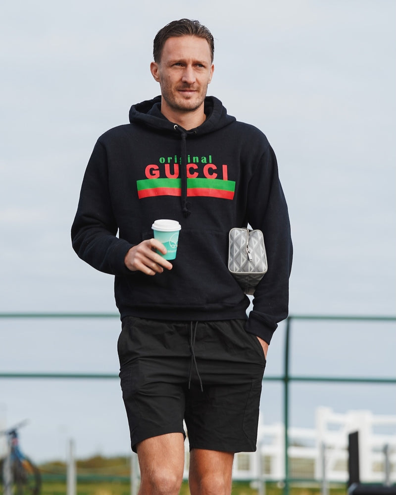Oxford United Defender, Benjamin Keith Davies, wearing a black hoodie with Gucci logo, holding a Missing Bean coffee cup and bag, standing outdoors.