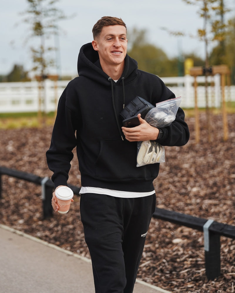 Oxford United Goalkeeper Jamie Cumming in black hoodie and trousers walking outdoors holding a Missing Bean coffee cup and bag.